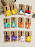 Twelve monster-themed luggage tags arranged on a light wood surface, each attached to a tan card with a black strap. The tags feature colorful character designs